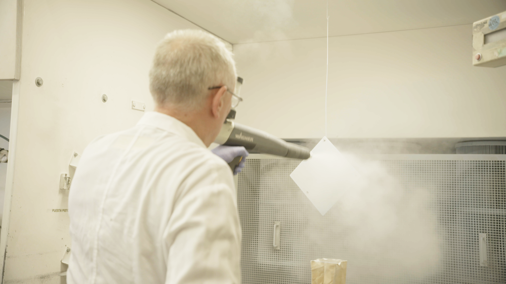 A man using an electrostatic spray gun to spray on enamel powder onto a square sheet of metal.